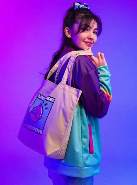 Tote Bag Tunisie 2 Sac en toile avec illustration originale, porté par une jeune femme souriante, look coloré et tendance, fond violet lumineux, style streetwear, mode casual dynamique, accessoire mode mode jeune, concept d'expression personnelle.