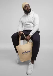 Stylé homme noir en sweat à capuche blanc, pantalon noir, chaussettes blanches, portant un sac en toile beige avec le mot "authentic" en lettres bleues, pose moderne, mode urbaine, style décontracté, vêtements casual, accessoires tendance, look streetwear, marque MarYouli®.